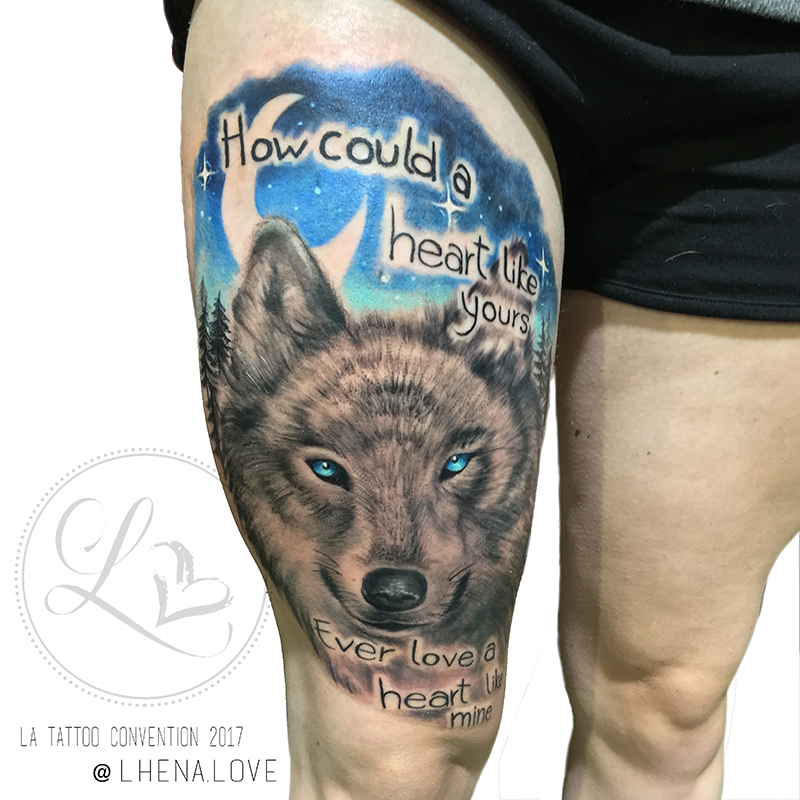 Black and grey realistic tattoo of a wolf with blue eyes against a blue night sky with moon and stars and lettering